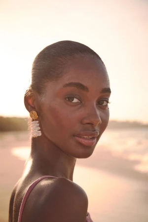 Model Tami Williams wearing layered pearl drop earrings and a pink top on a beach at sunset.