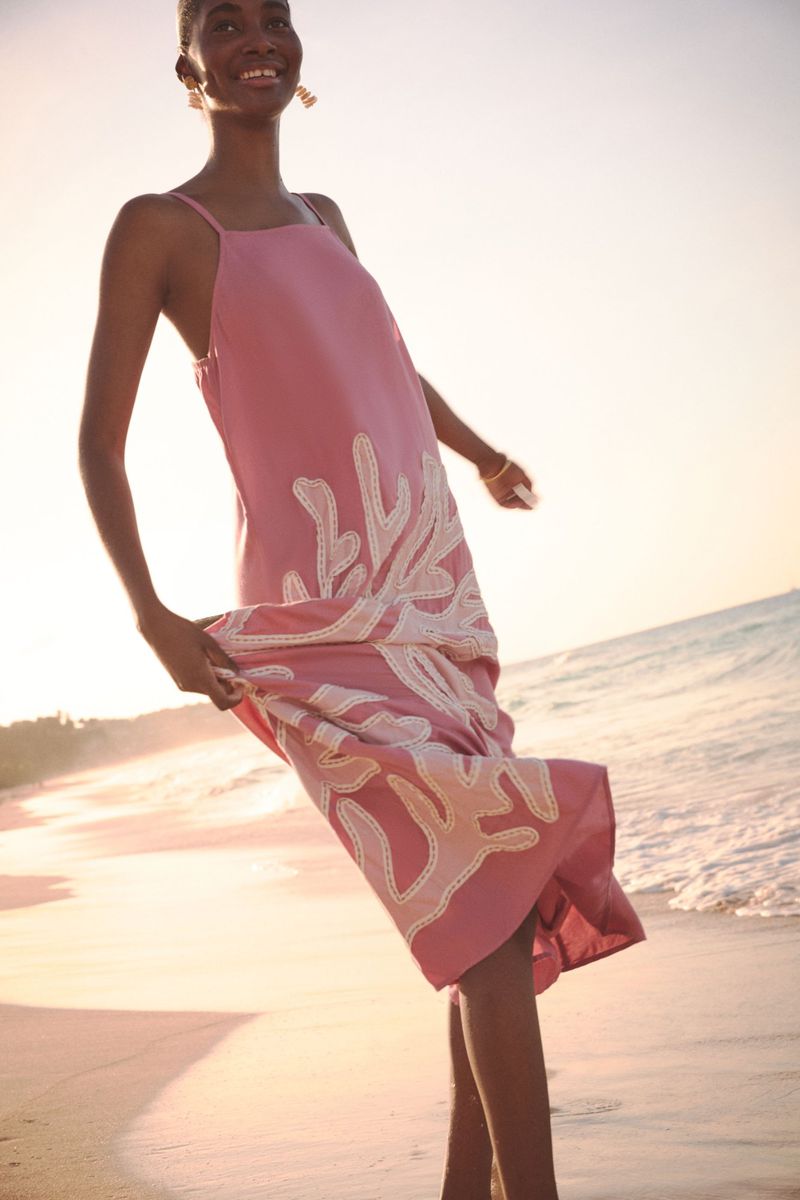 A smiling black woman in a pink coral-patterned dress stands on a beach with golden sand and ocean waves in the background in sunlight.