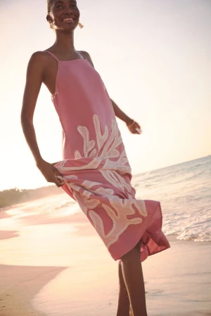 A smiling black woman in a pink coral-patterned dress stands on a beach with golden sand and ocean waves in the background in sunlight.