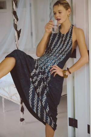 Model Paige Reifler wearing a navy patterned halter midi dress and holding a glass of water, leaning against a white doorway.