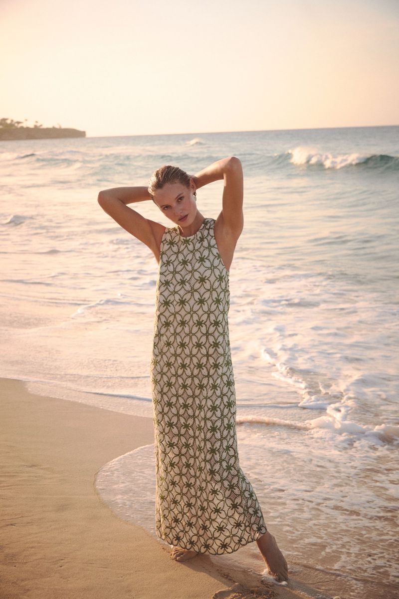 Model in a patterned green and cream maxi dress posing barefoot on a sandy beach with ocean surf in the background.