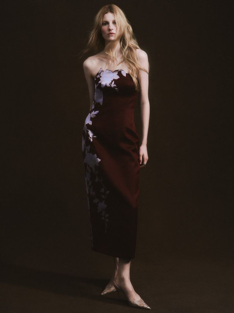 Woman poses in a strapless wine-colored dress with floral detail against a dark brown background in a studio fashion shot.