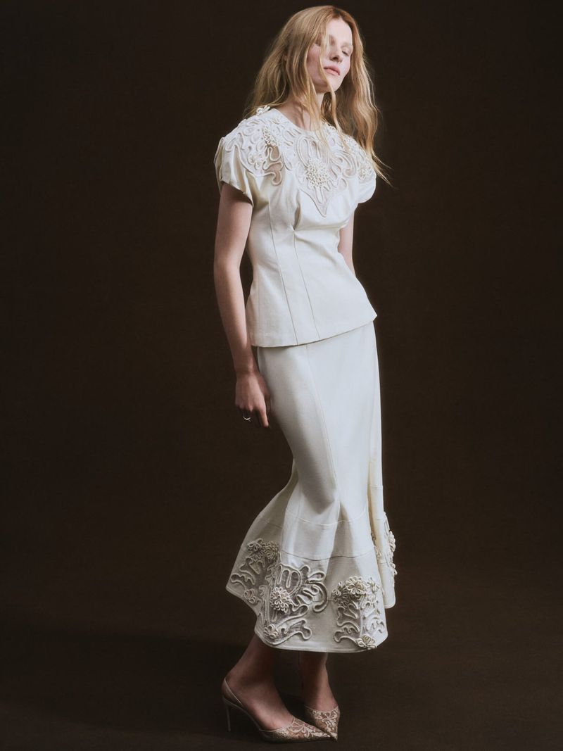 A full shot of a blonde adult female model in a light-beige embroidered blouse and a long skirt, paired with light, stiletto heels.