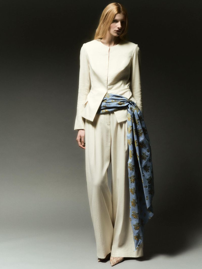 Fashionable woman in cream-colored jacket and palazzo pants, styled with an ornate blue sash, poses in a studio setting.