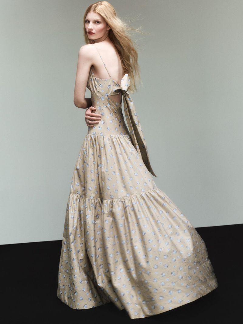 Elegant blonde woman posing in a long, floral-patterned dress with a large bow. Fashion shot with a neutral studio backdrop.