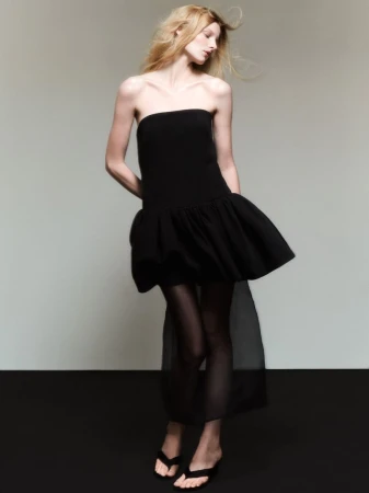 Model in a black strapless mini dress standing against a plain grey studio backdrop for Lela Rose lookbook