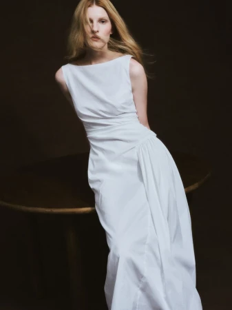 Model with long blonde hair wearing a sleeveless white dress leaning against a dark wooden table in studio.