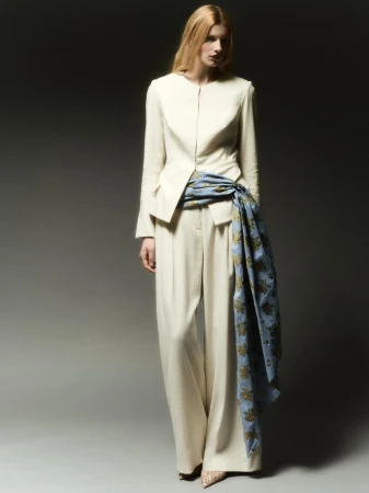 Model standing on a grey backdrop wearing a cream jacket and trousers with a blue patterned scarf tied around the waist.