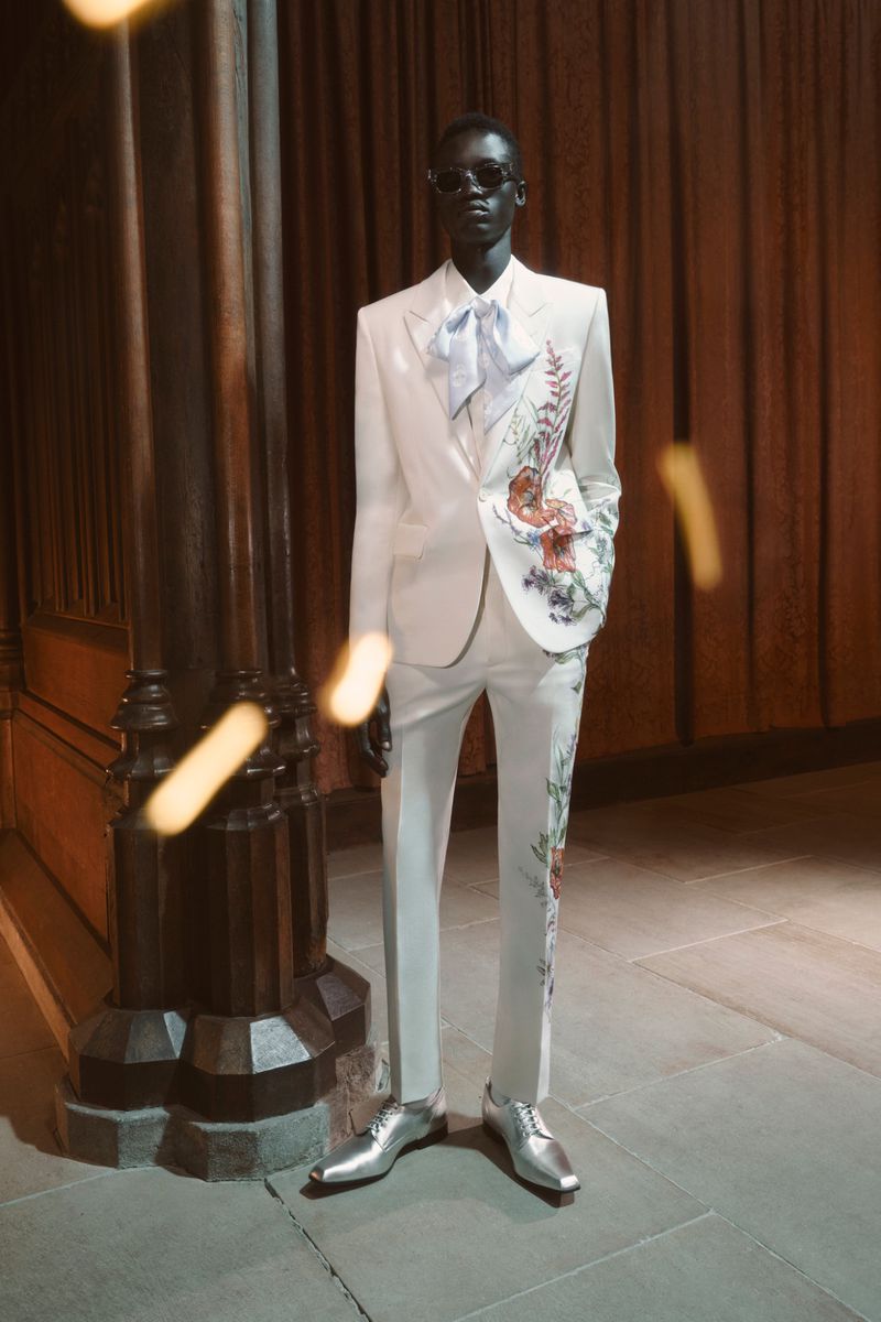A dark-skinned male fashion model poses in a white floral-patterned suit with shiny silver shoes, standing indoors with a neutral expression.