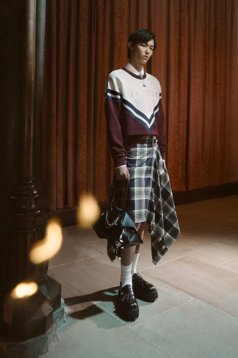An adult fashion model stands in a stylish outfit consisting of a sweater, asymmetrical plaid skirt, chunky loafers and a purse.
