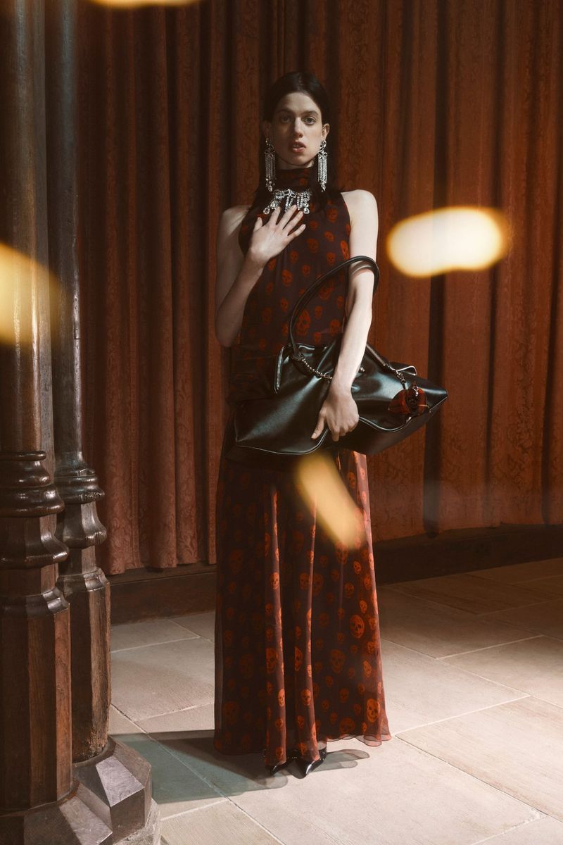A fashionable woman in a long dress and statement jewelry stands in front of a curtain, holding a large black bag with a flower.