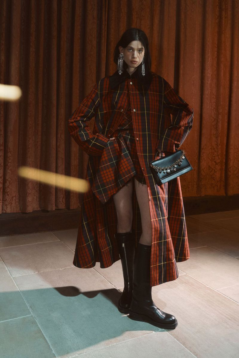 A woman in a red and green plaid coat and black boots poses in front of a red curtain, holding a black embellished purse.