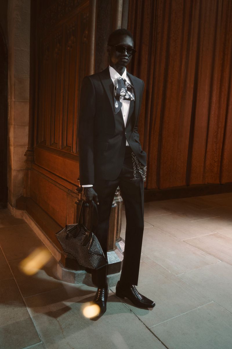 A slender black man in a black suit and black sunglasses stands in front of dark wood panelling, holding a dark patterned bag.