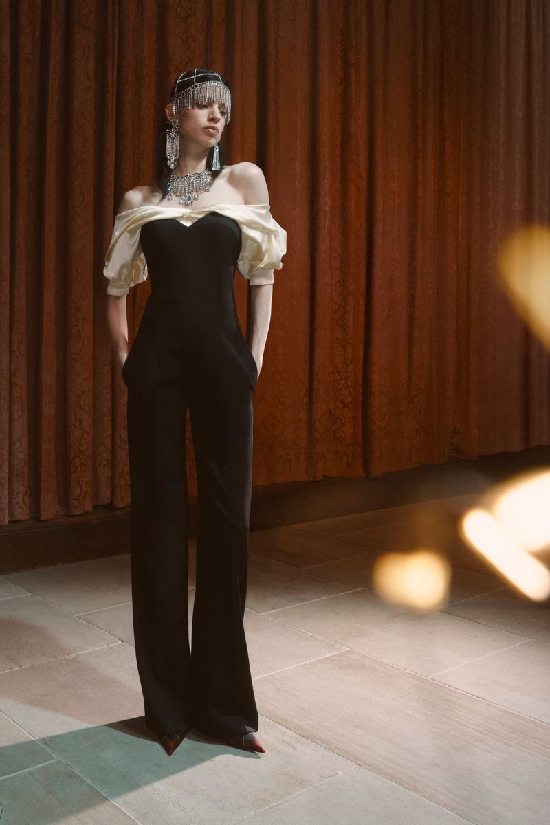 Elegant woman poses in a black jumpsuit with off-the-shoulder cream sleeves, complemented by a jewel headpiece and statement jewelry indoors.