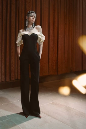 Elegant woman poses in a black jumpsuit with off-the-shoulder cream sleeves, complemented by a jewel headpiece and statement jewelry indoors.