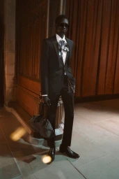 Model Jum Kuochnin in a black tailored suit, sunglasses, and printed necktie holding a tote bag in a wood-paneled interior.