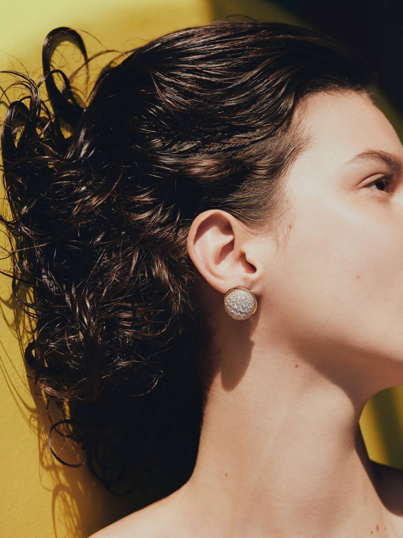 Close-up studio portrait of a brunette adult female wearing a round, decorative earring against a vibrant yellow backdrop.