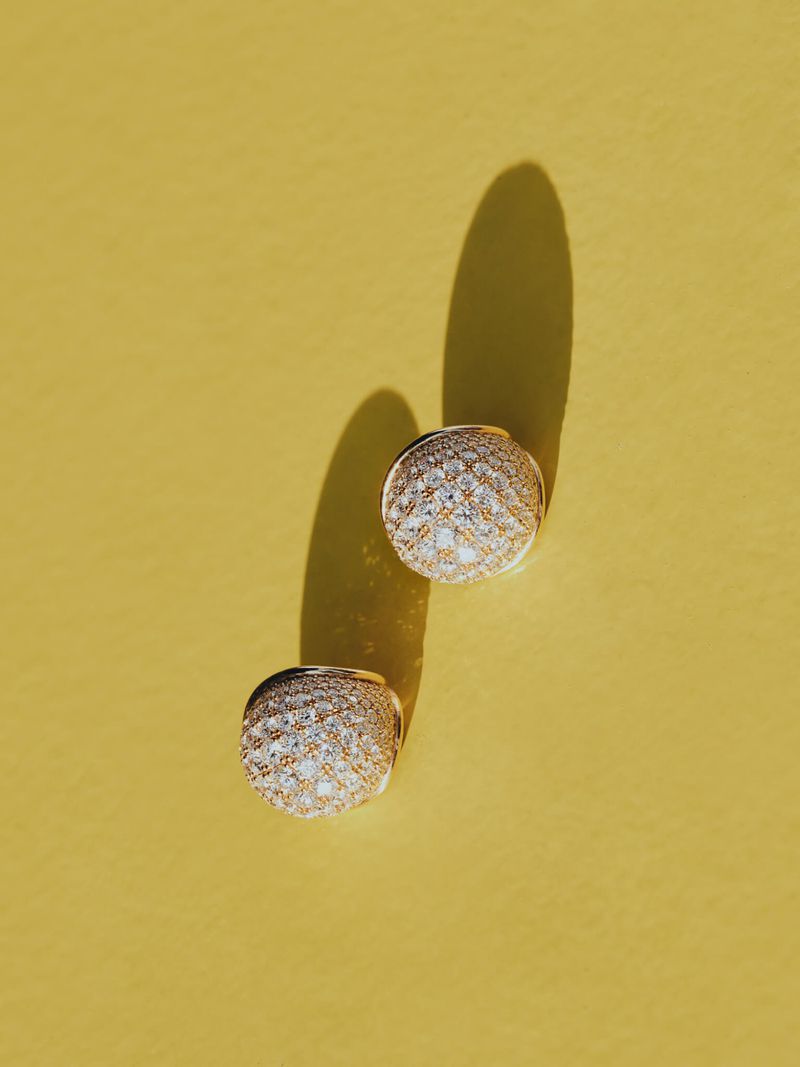 Overhead shot of two gold and diamond stud earrings on a sunny yellow background, casting long shadows with soft light.