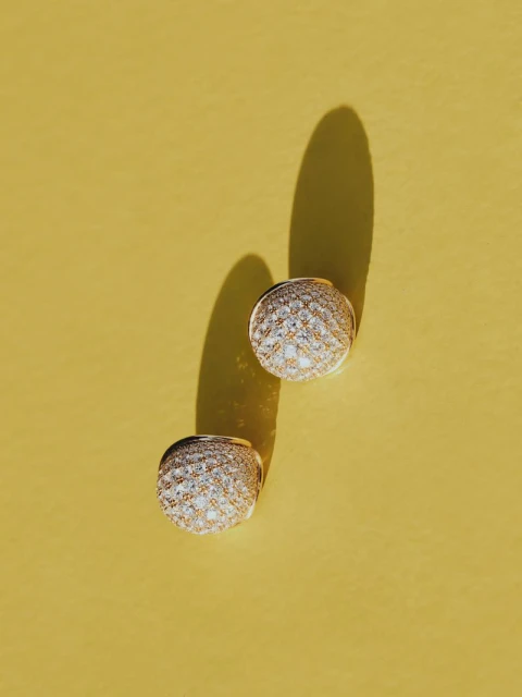 Overhead shot of two gold and diamond stud earrings on a sunny yellow background, casting long shadows with soft light.
