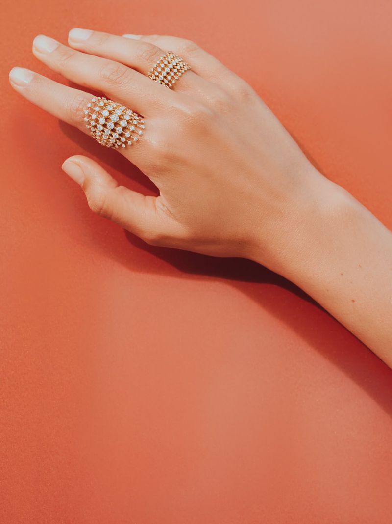 Close-up of a hand with fair skin and two gold lattice-style rings with small stones, set against a coral-colored background. Beauty shot.
