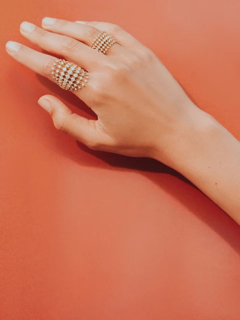Close-up of a hand with fair skin and two gold lattice-style rings with small stones, set against a coral-colored background. Beauty shot.