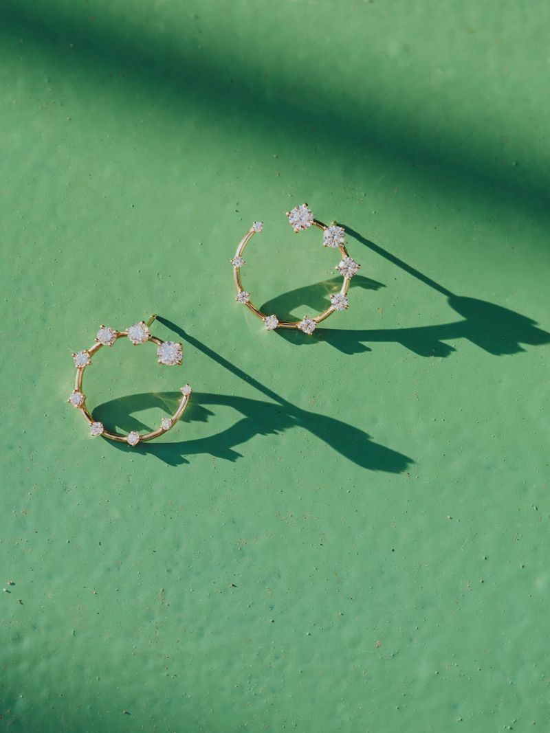 Close-up of gold hoop earrings adorned with gemstones on a textured green surface, illuminated by dappled sunlight, casting sharp shadows.