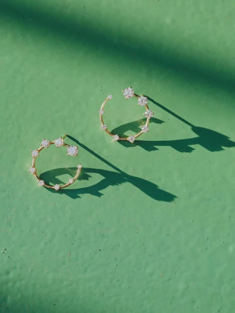 Close-up of gold hoop earrings adorned with gemstones on a textured green surface, illuminated by dappled sunlight, casting sharp shadows.