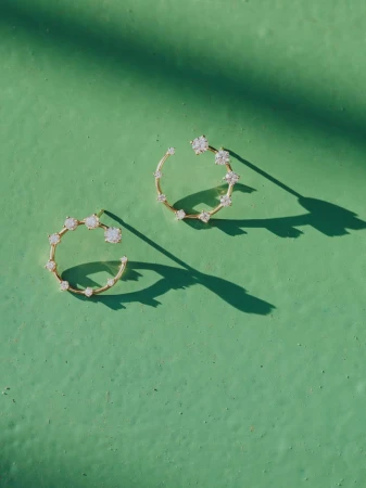 Close-up of gold hoop earrings adorned with gemstones on a textured green surface, illuminated by dappled sunlight, casting sharp shadows.