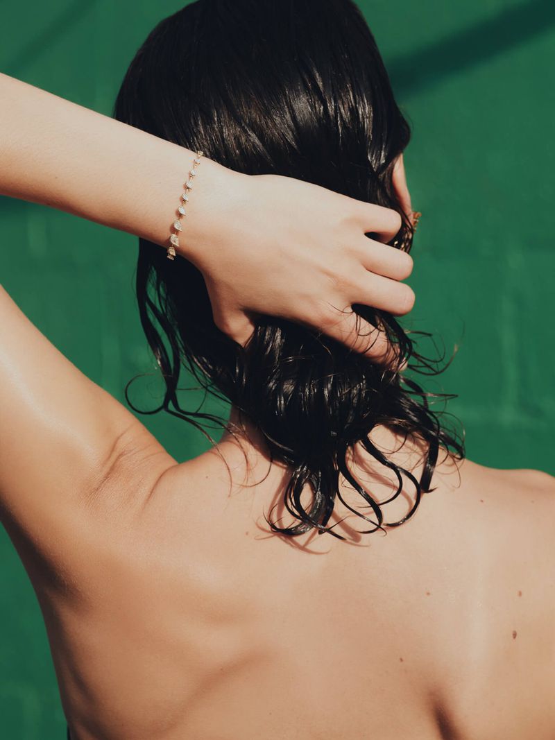 Back view of an adult woman with wet dark hair wearing a bracelet against a solid green background in a studio setting.