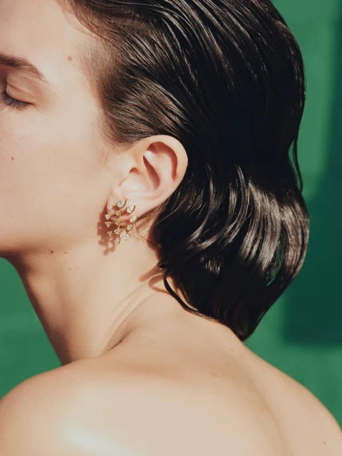 Close-up studio shot of an adult woman with wet black hair wearing floral earrings, against a green background with diffused lighting.