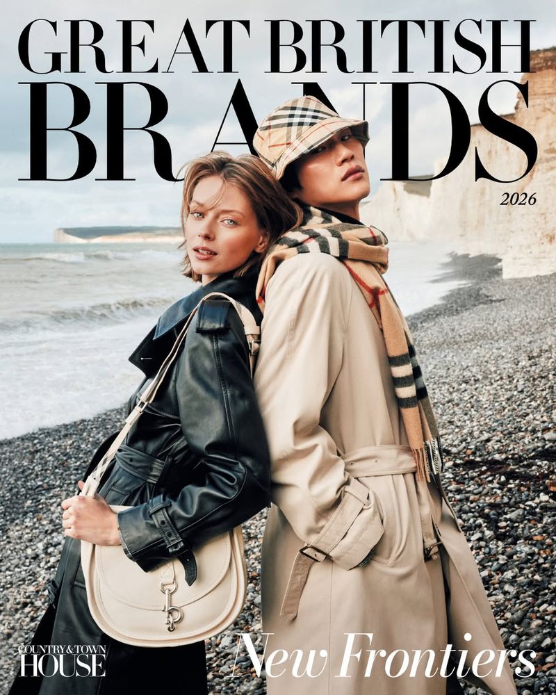 Two fashion models pose back to back on a rocky beach in tan trench coats with the ocean and cliffs in the background in editorial fashion shoot.