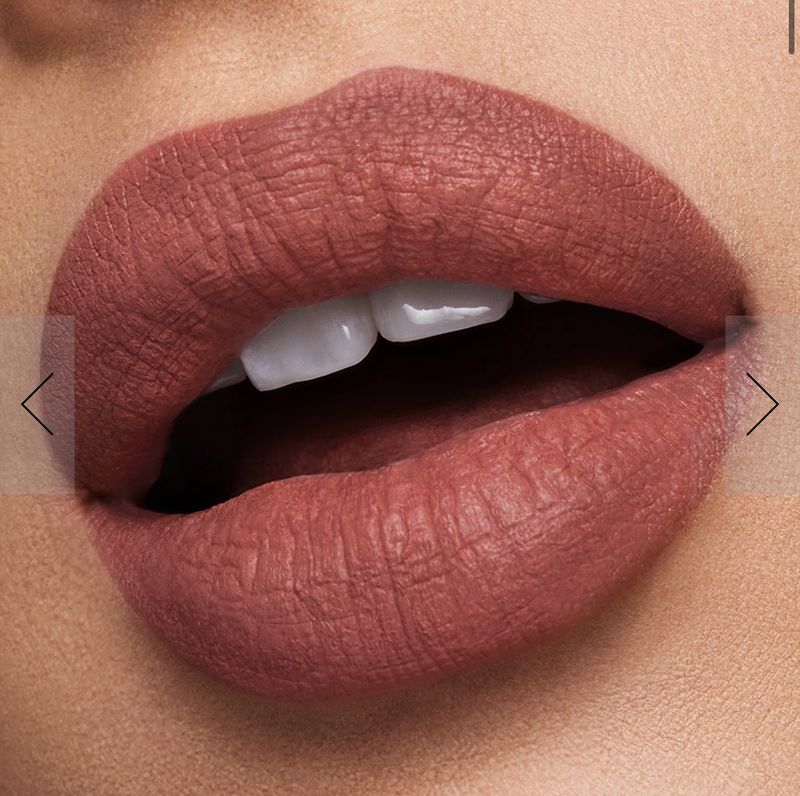 Close-up of open mouth with even white teeth and lips covered in matte brownish-red lipstick against a light skin-tone background.