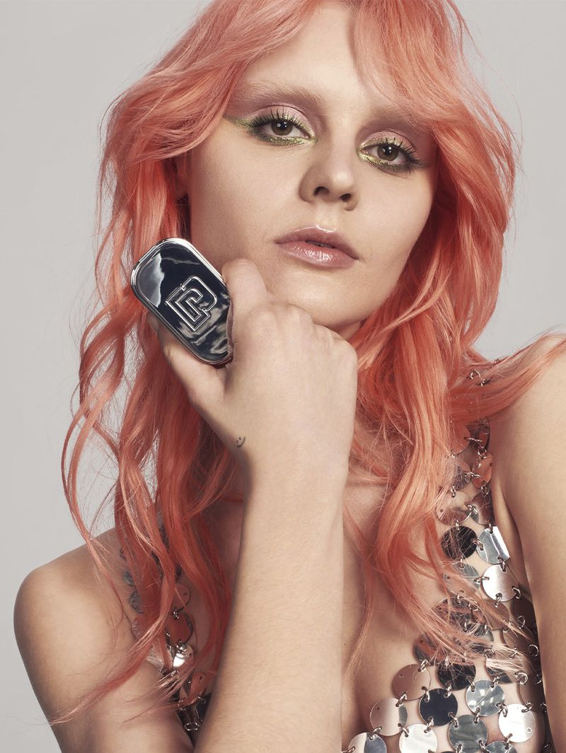 Close-up portrait of a woman with pink hair and shimmering makeup wearing a silver sequined top against a neutral grey backdrop.