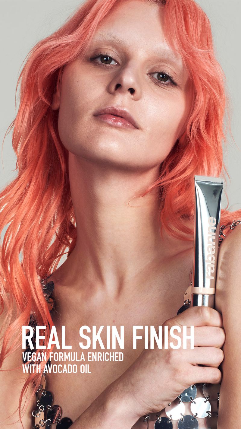 Woman with orange hair holds a silver tube of RABANNE Real Skin Finish vegan formula makeup. Close-up studio shot against a neutral background.