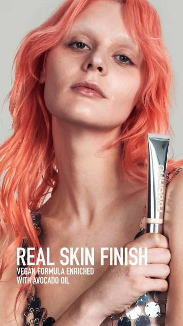 Woman with orange hair holds a silver tube of RABANNE Real Skin Finish vegan formula makeup. Close-up studio shot against a neutral background.