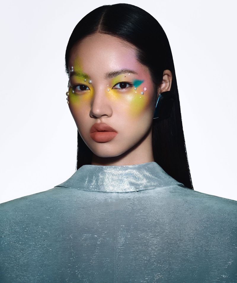 Close-up of a woman with creative yellow, pink, and teal eye makeup, glossy skin, and a shiny collared shirt against a white backdrop.
