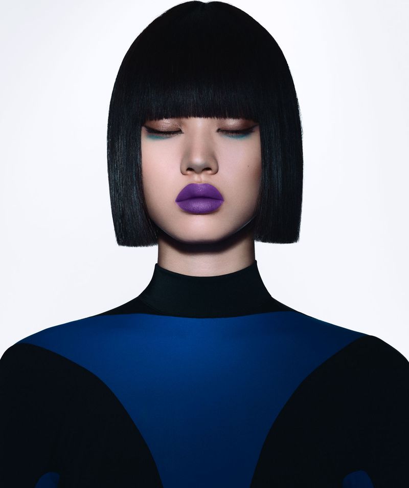 Close-up portrait of an Asian woman with a short, blunt black bob, wearing blue eye shadow, purple lipstick, and a black and blue top.