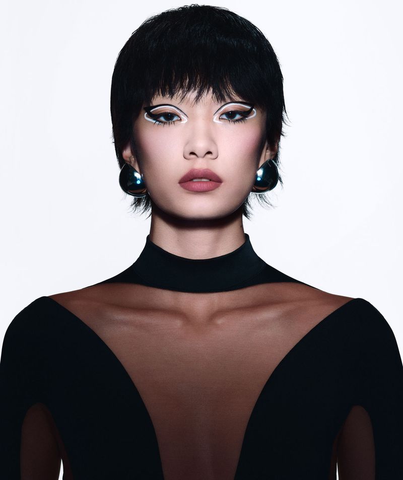 Close-up of an Asian woman with short black hair, striking white eyeliner, and a black top with a sheer neckline, posing in a studio.