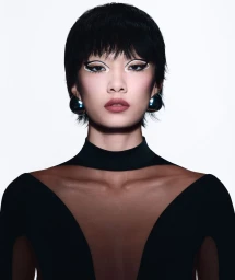 Close-up portrait of a model with short dark hair, graphic white eyeliner, and large metallic drop earrings against a white background.