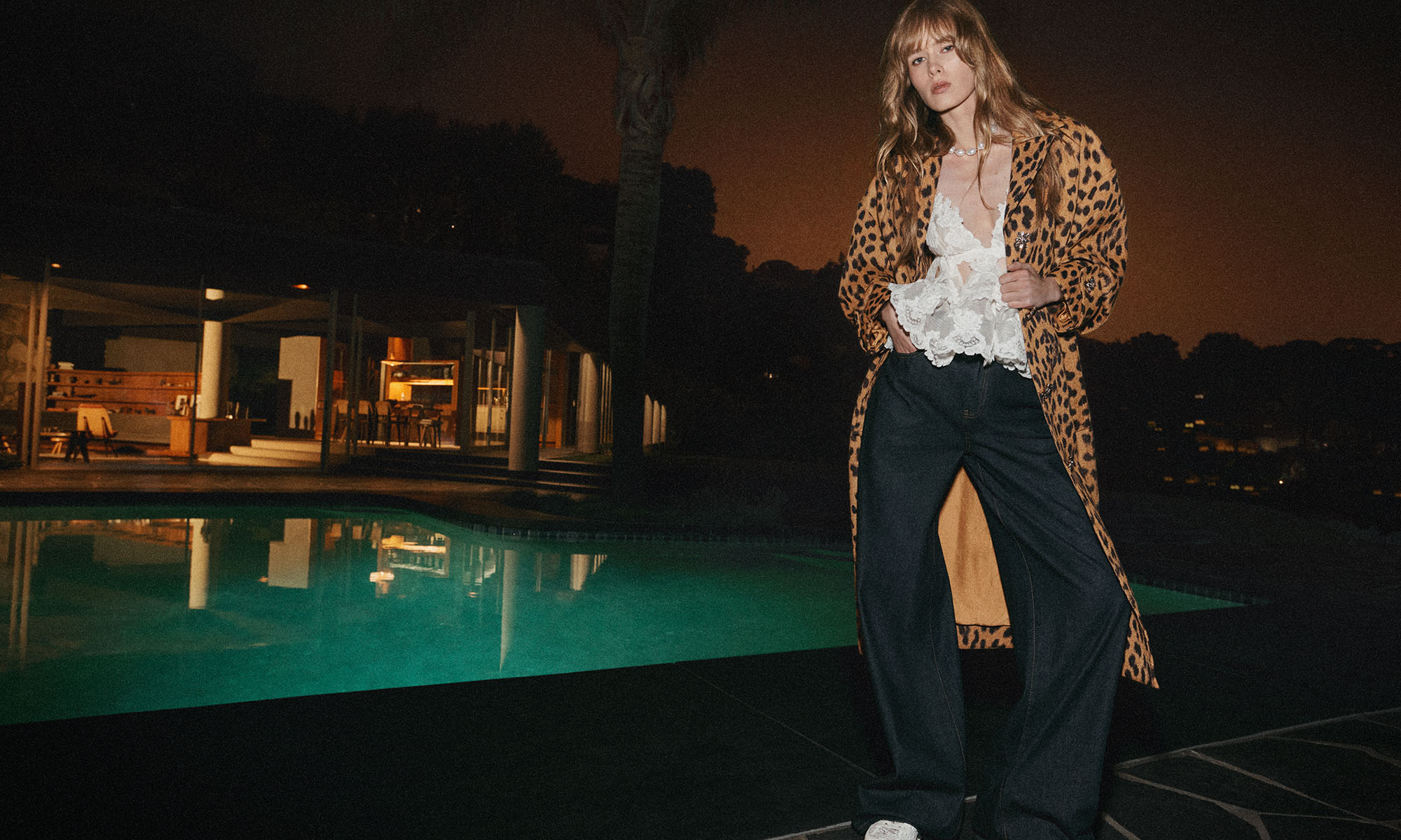 Woman in animal-print coat poses poolside at night. She wears a lace top and dark wide-leg jeans, with modern home in background.