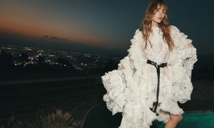 Model in ruffled white lace dress with a black belt posing outdoors at dusk against a city skyline view