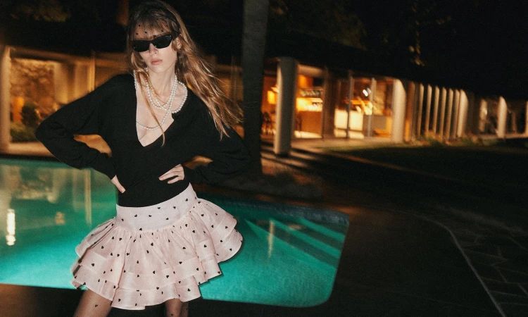 Model wearing black V-neck top, pink ruffled mini skirt, pearl necklaces, and sunglasses by a glowing outdoor swimming pool at night.