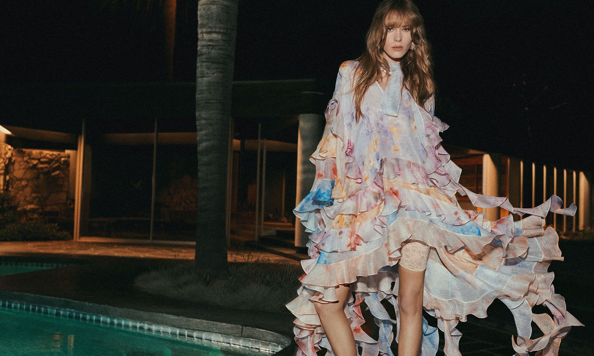 A lone female model in a ruffled pastel dress stands beside a pool at night, with a modern house and palm tree visible in the background.