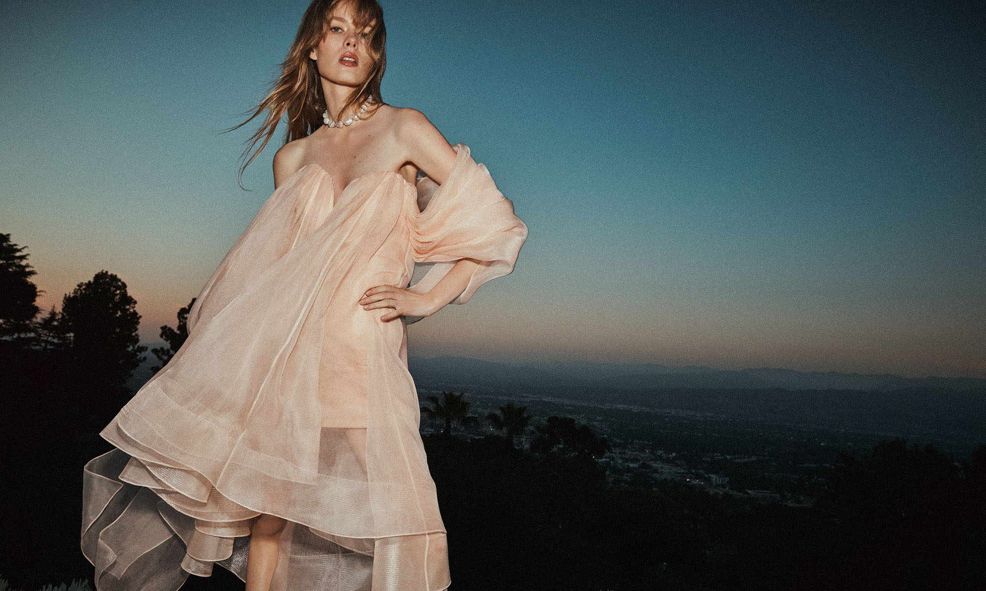 A woman in a light pink dress and pearl necklace poses outdoors at dusk, with a city and distant mountains in the background.