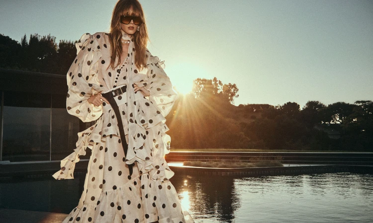 An adult woman in a polka dot dress poses with her hands on her hips in front of a home, trees, and water at sunset.