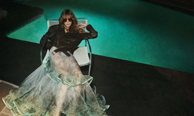Woman in dark sunglasses wearing a black leather jacket and patterned gown seated by a glowing teal swimming pool at night.