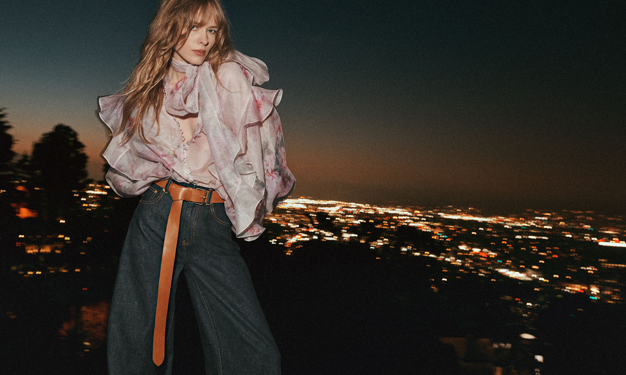 A woman poses in a semi-sheer floral blouse and jeans, cinched with a brown leather belt, set against an urban nightscape.