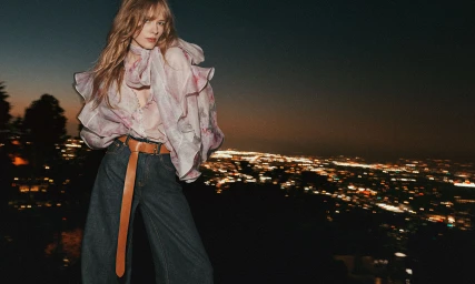 Model in a floral ruffled sheer blouse and wide-leg jeans with a long tan belt against a night city skyline backdrop.