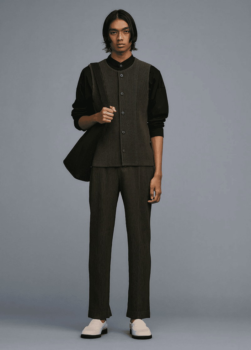 Full-length shot of an adult male model in a grey photo studio wearing pleated olive-green pants, vest, black shirt, white shoes, and carrying a shoulder bag.