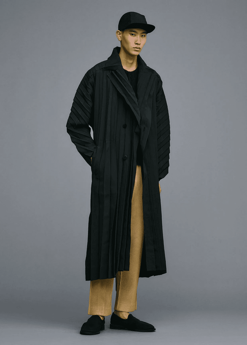 Asian male fashion model posing in a pleated black coat and pants, in a studio with grey background.